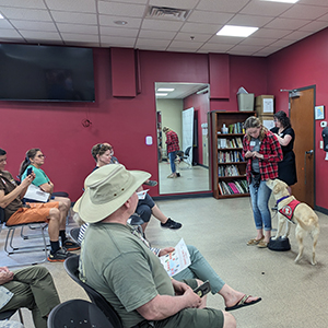 woman standing at front of room of people with yellow Lab service dog standing with front legs on perch looking up at her