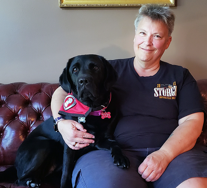 woman sitting on leather sofa with arm around black Lab service dog lying next to her