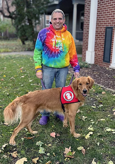 man standing on grass outside building wearing colorful tie-dyed sweatshirt and Golden Retriever wearing red Can Do Canines service dog cape; both are smiling at camera