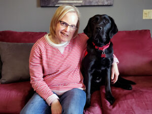 woman sitting on red sofa with arm around black Lab dog sitting next to her; both are smiling at camera