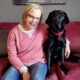 woman sitting on red sofa with arm around black Lab dog sitting next to her; both are smiling at camera