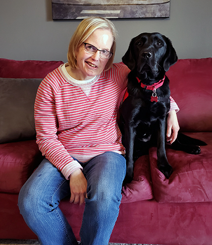 woman sitting on red sofa with arm around black Lab dog sitting next to her; both are smiling at camera