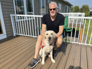 man crouching on deck with yellow Lab service dog sitting between his legs; both are looking at camera