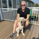 man crouching on deck with yellow Lab service dog sitting between his legs; both are looking at camera