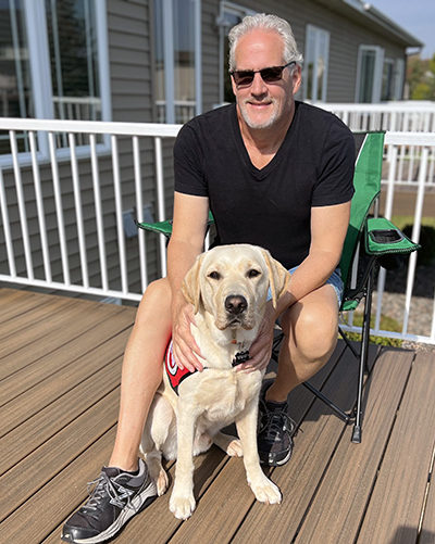 man crouching on deck with yellow Lab service dog sitting between his legs; both are looking at camera