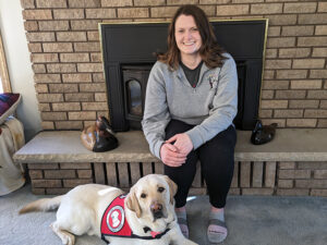 woman sitting on fireplace hearth with yellow Lab service dog wearing red Can Do Canines cape at her feet