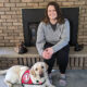 woman sitting on fireplace hearth with yellow Lab service dog wearing red Can Do Canines cape at her feet