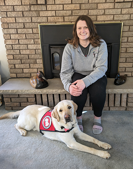 woman sitting on fireplace hearth with yellow Lab service dog wearing red Can Do Canines cape at her feet