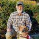 man with beard and cap crouching on grass with hand on yellow Lab that is wearing red service cape; both are looking at camera