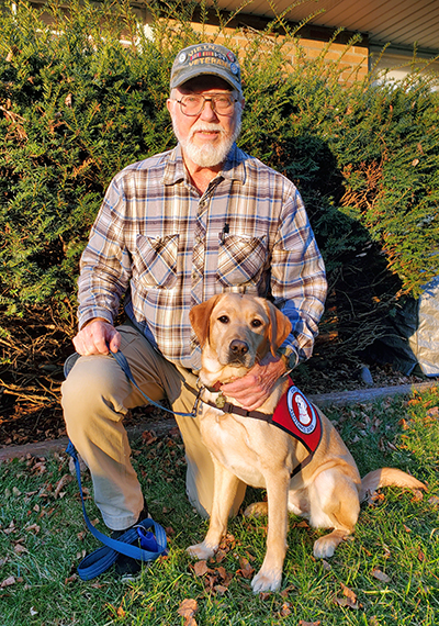 man with beard and cap crouching on grass with hand on yellow Lab that is wearing red service cape; both are looking at camera