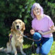 woman crouching on grass next to Golden Retriever that is wearing a red service cape