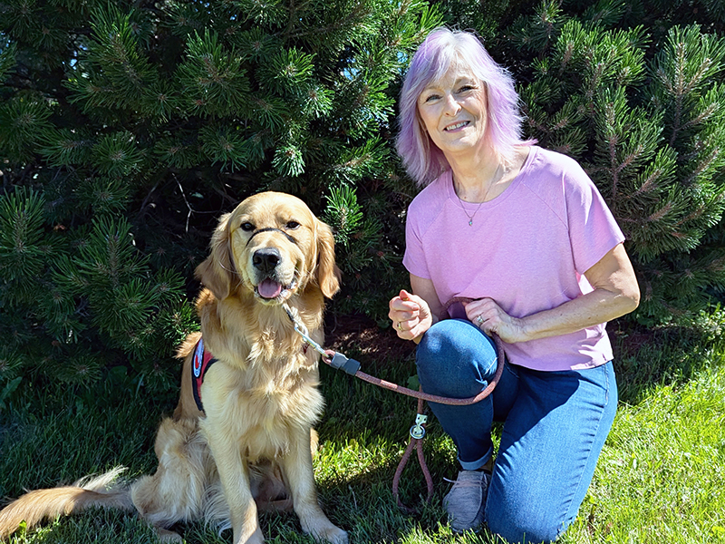 woman crouching on grass next to Golden Retriever that is wearing a red service cape