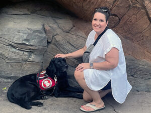 woman crouching down on rock formation to put hand on black Lab service dog wearing Can Do Canines cape; both are looking at camera