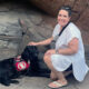 woman crouching down on rock formation to put hand on black Lab service dog wearing Can Do Canines cape; both are looking at camera