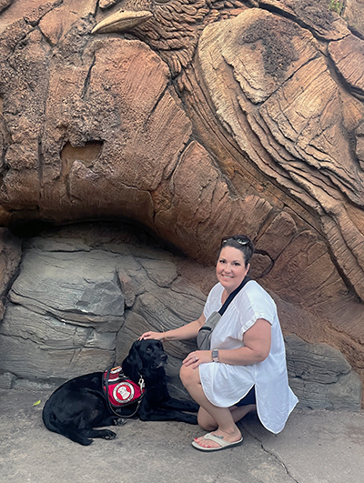 woman crouching down on rock formation to put hand on black Lab service dog wearing Can Do Canines cape; both are looking at camera