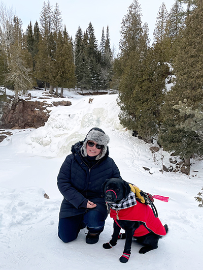 woman crouching down on snow near pine trees to put hand on black Lab service dog wearing red coat; both are looking at camera