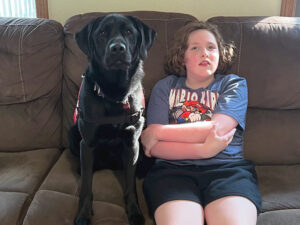 girl sitting on sofa next to black Lab service dog, both are looking at camera