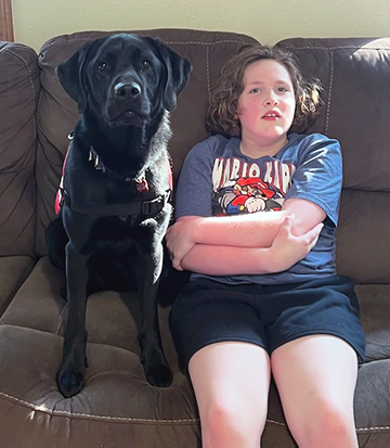 girl sitting on sofa next to black Lab service dog, both are looking at camera