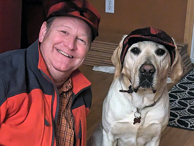 man wearing red and black jacket and red and black plaid hat, sitting on floor next to yellow Lab with matching hat, both are smiling at camera