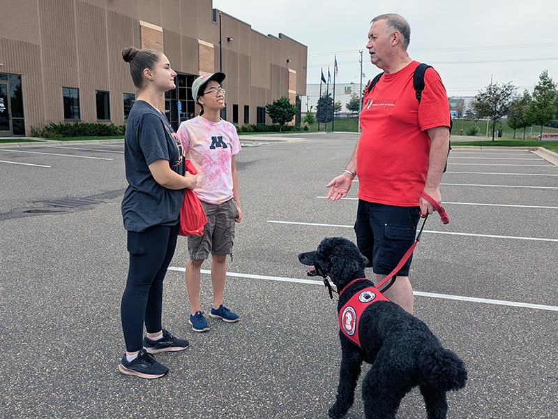 man with black Standard Poodle service dog standing in parking lot talking with two young women