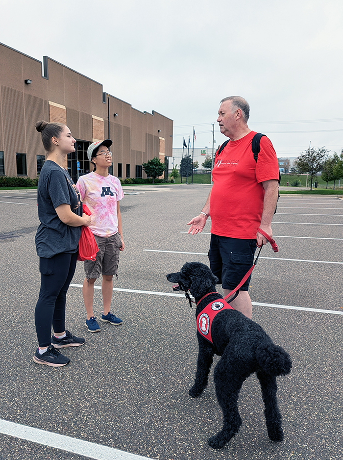 man with black Standard Poodle service dog standing in parking lot talking with two young women