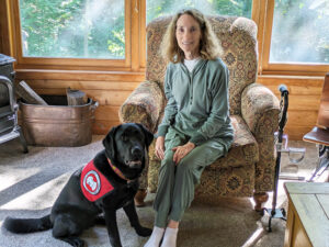 woman sitting in upholstered cair with black Lab service dog wearing red Can Do Canines cape sitting near her legs; both are looking at the camera