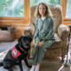 woman sitting in upholstered cair with black Lab service dog wearing red Can Do Canines cape sitting near her legs; both are looking at the camera