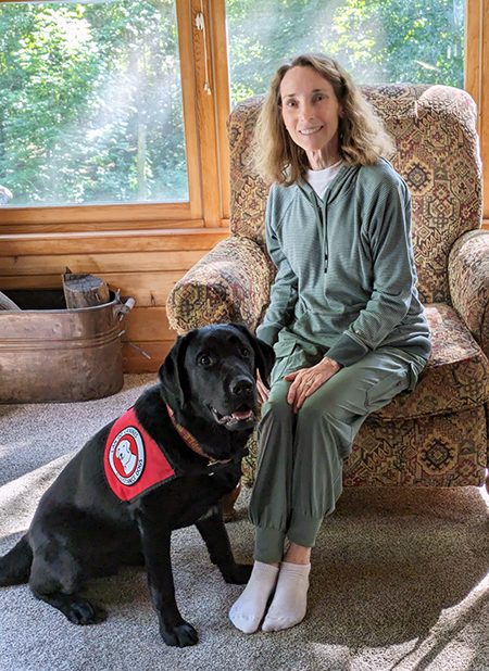 woman sitting in upholstered cair with black Lab service dog wearing red Can Do Canines cape sitting near her legs; both are looking at the camera