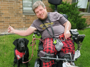 man sitting outside in power chair next to black Lab service dog, both are smiling at camera