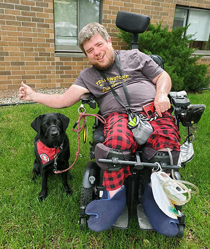 man sitting outside in power chair next to black Lab service dog, both are smiling at camera