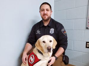 male police officer crouching on floor with yellow Lab service dog wearing Can Do Canines cape sitting in front of him; both are looking at camera