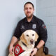 male police officer crouching on floor with yellow Lab service dog wearing Can Do Canines cape sitting in front of him; both are looking at camera