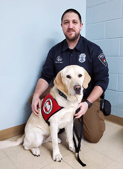 male police officer crouching on floor with yellow Lab service dog wearing Can Do Canines cape sitting in front of him; both are looking at camera