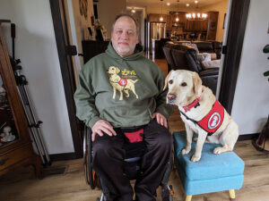 man sitting in wheelchair in home with yellow Lab service dog wearing red Can Do Canines cape sitting on ottoman next to him; both are looking at camera