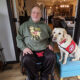 man sitting in wheelchair in home with yellow Lab service dog wearing red Can Do Canines cape sitting on ottoman next to him; both are looking at camera