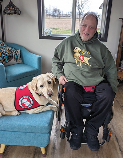 man sitting in wheelchair in home with yellow Lab service dog wearing red Can Do Canines cape sitting on ottoman next to him; both are looking at camera