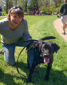 woman kneels in the grass next to a black Lab.