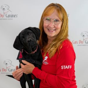 Woman wearing a red Can Do Canines staff shirt is holding a young black Lab puppy and smiling at the camera.