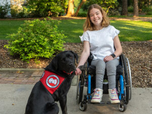 A teenage girl using a wheelchair is smiling at the camera and her black Labrador service dog is sitting next to her with his paw placed in her hand.