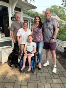 A teenage girl using a wheelchair is smiling at the camera and her black Labrador service dog is sitting next to her. Surroung the girl and dog, are two couples.