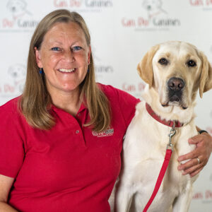 A woman wearing a red Can Do Canines staff shirt poses with a yellow Labrador. 