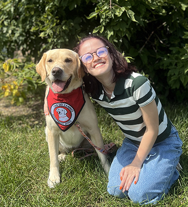 young woman sitting on grass with arm around yellow Lab service dog wearing red scarf; both are smiling at the camera