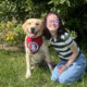 young woman sitting on grass with arm around yellow Lab service dog wearing red scarf; both are smiling at the camera