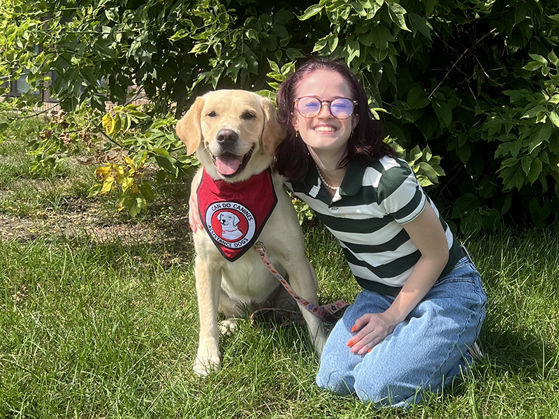 young woman sitting on grass with arm around yellow Lab service dog wearing red scarf; both are smiling at the camera