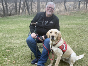 man sitting on chair on grass next to yellow Lab service dog wearing red cape; both are smiling at the camera