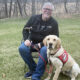 man sitting on chair on grass next to yellow Lab service dog wearing red cape; both are smiling at the camera