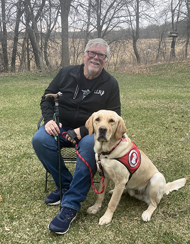 man sitting on chair on grass next to yellow Lab service dog wearing red cape; both are smiling at the camera