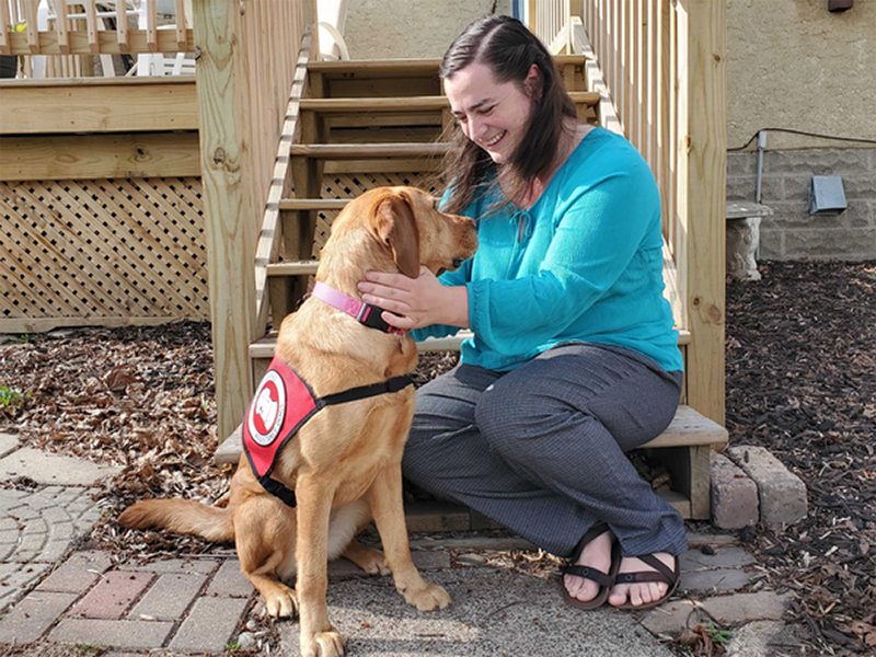 woman sitting on deck steps smiling at and putting hands on yellow Lab service dog wearing red cape