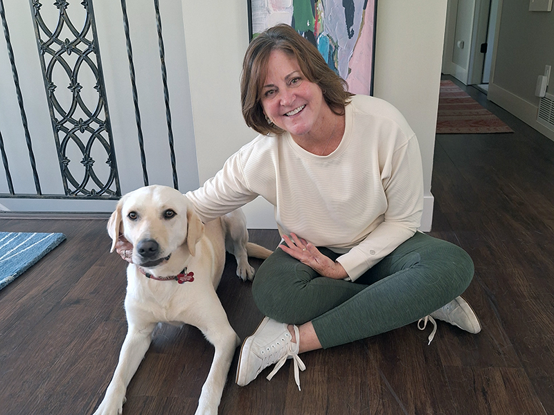 woman sitting on hardwood floor with hand on yellow Lab; both are smiling at the camera