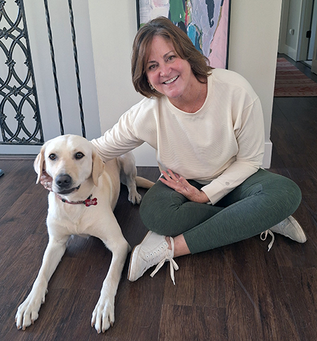 woman sitting on hardwood floor with hand on yellow Lab; both are smiling at the camera